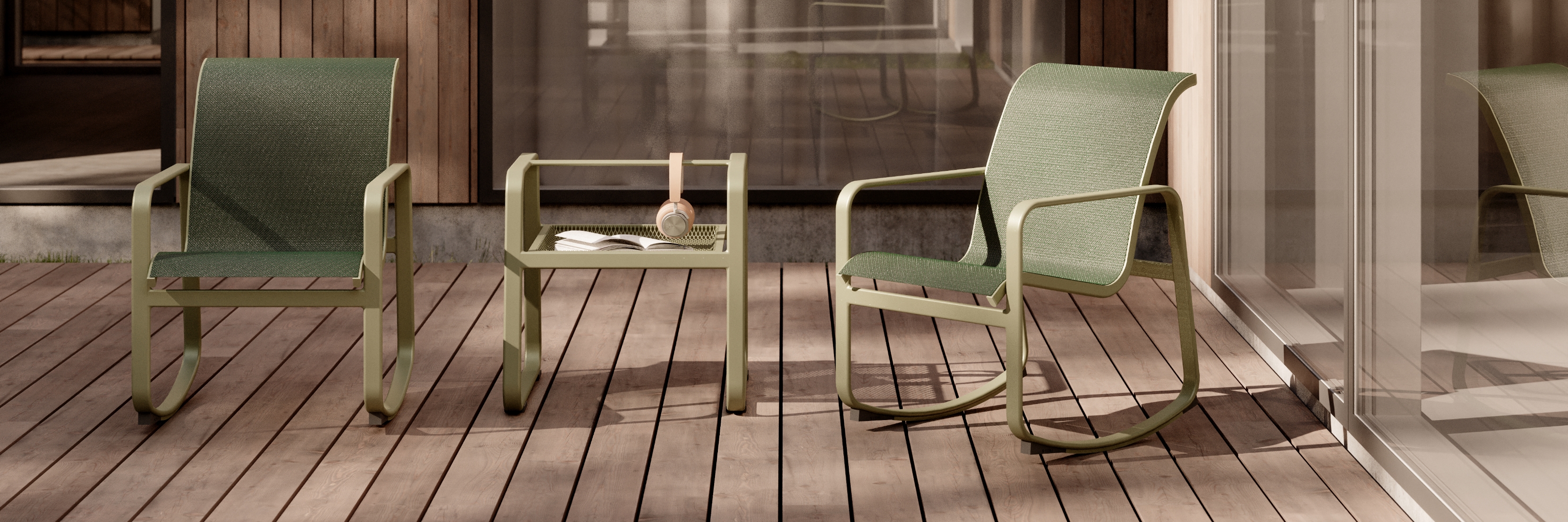Two green rocking chairs from the Sequel collection flank a matching table on a wooden deck. The table holds a book and a teacup. The setting is outdoors, next to a glass wall and wooden paneling, creating a serene and cozy atmosphere.
